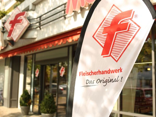 Werbefahne mit dem Logo des Fleischerhandwerks vor einem Fleischerfachgeschäft. Das Bild verweist auf die Dienstleistungen des Fleischerhandwerks.